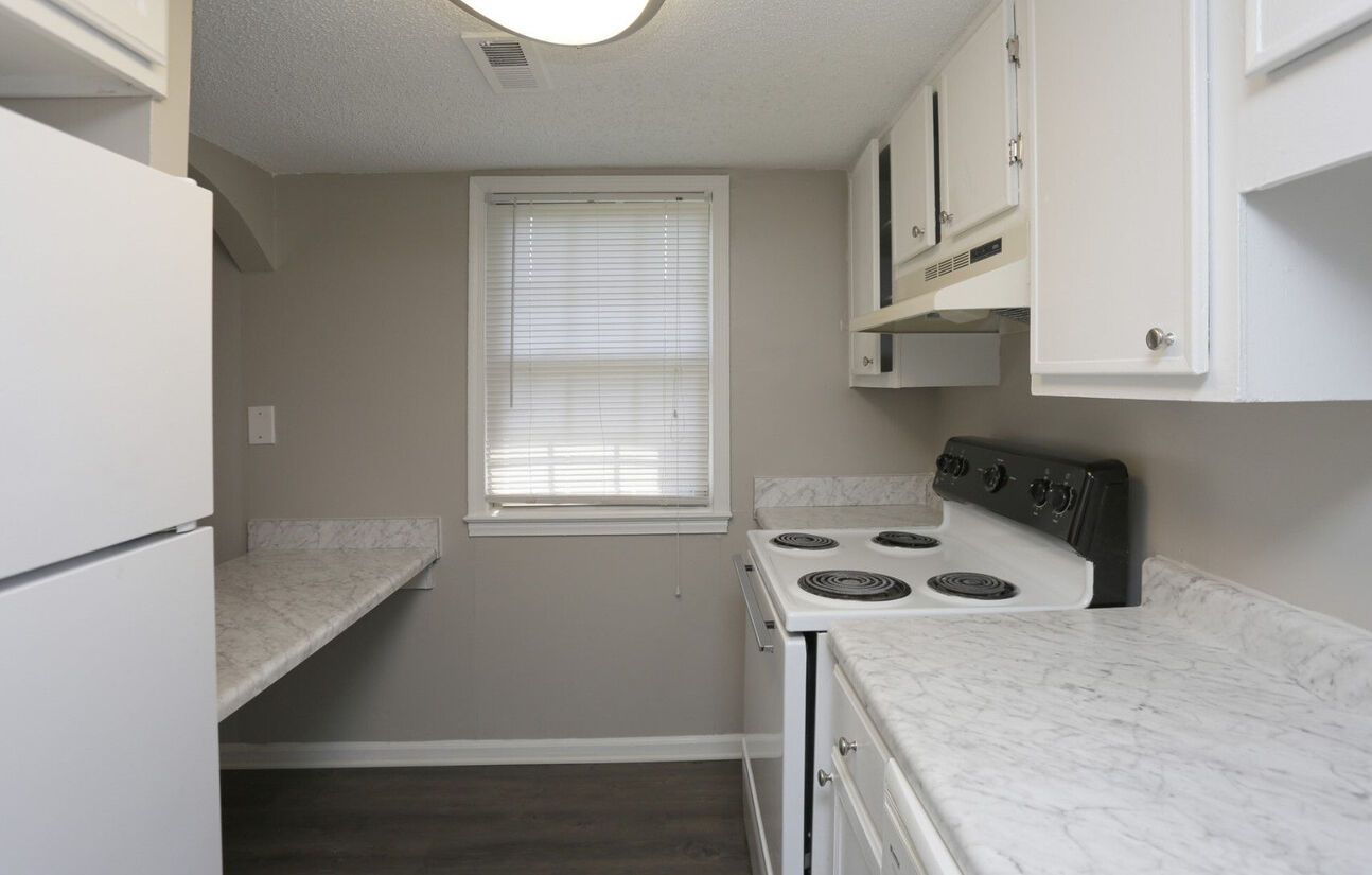 Small, white kitchen with light countertops and appliances. Window with blinds and a stovetop on the right.