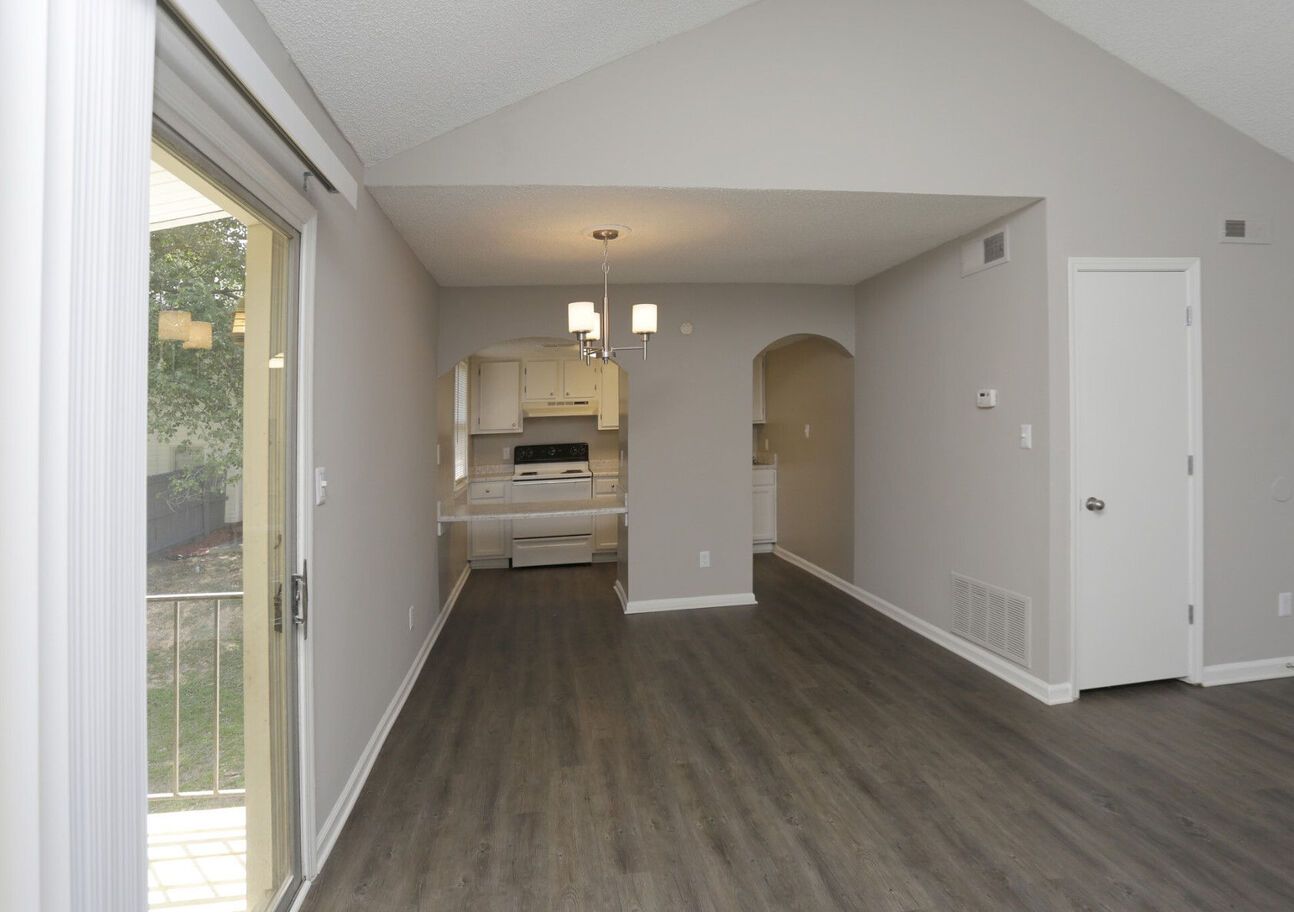 Interior view of a modern living space with gray walls, wood-look flooring, and a view into the kitchen. A sliding glass door leads to a balcony.