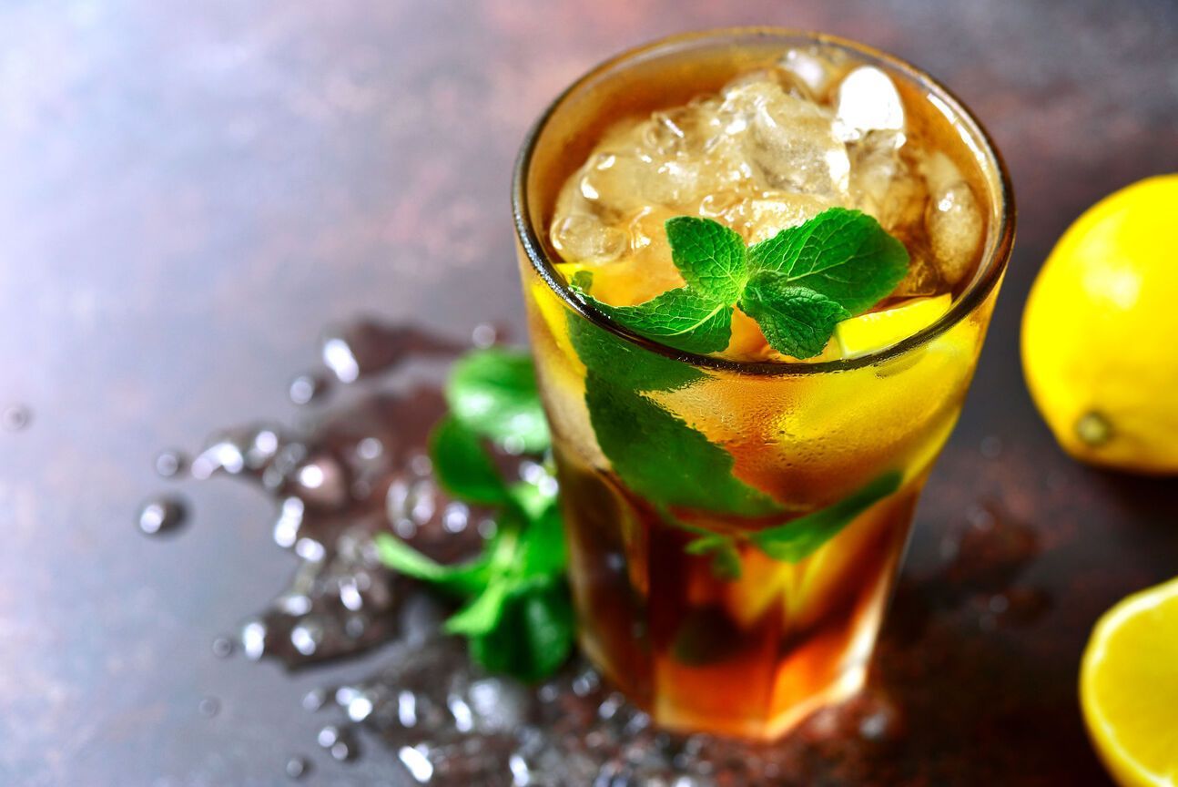 Iced tea with lemon slices, mint sprigs, and ice in a tall glass on a dark, wet surface; a whole and halved lemon are visible nearby.