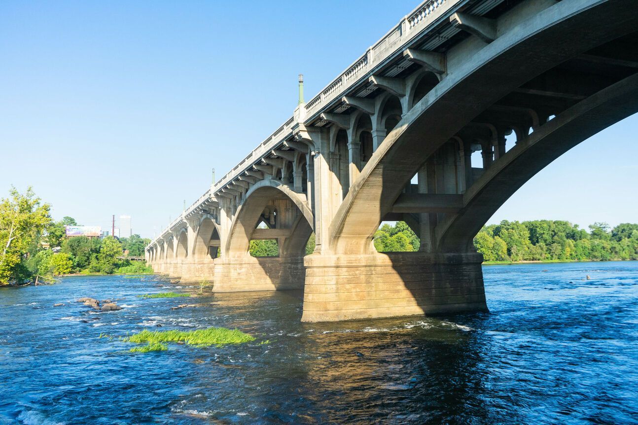 Arched concrete bridge spans a wide, blue river under a clear sky. Lush green trees line the riverbank.