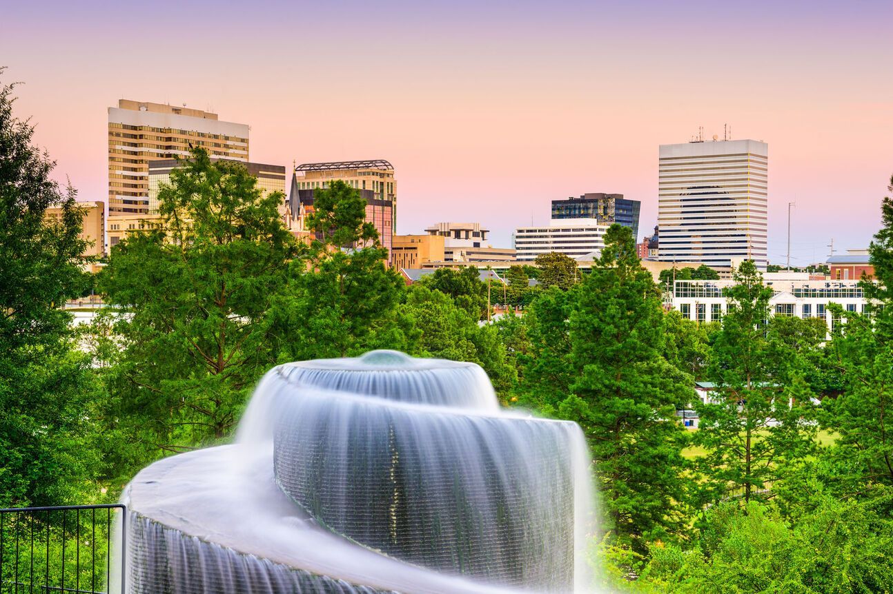 A scenic view of a city skyline with a flowing tiered fountain in the foreground and lush green trees. The sky is pastel pink and purple.