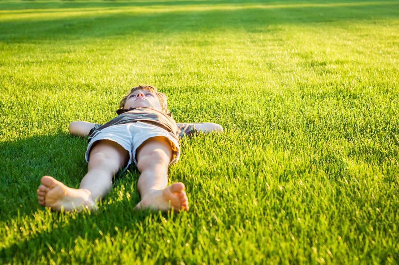 Child lying in green grass, arms outstretched, eyes closed, enjoying the sunlight.