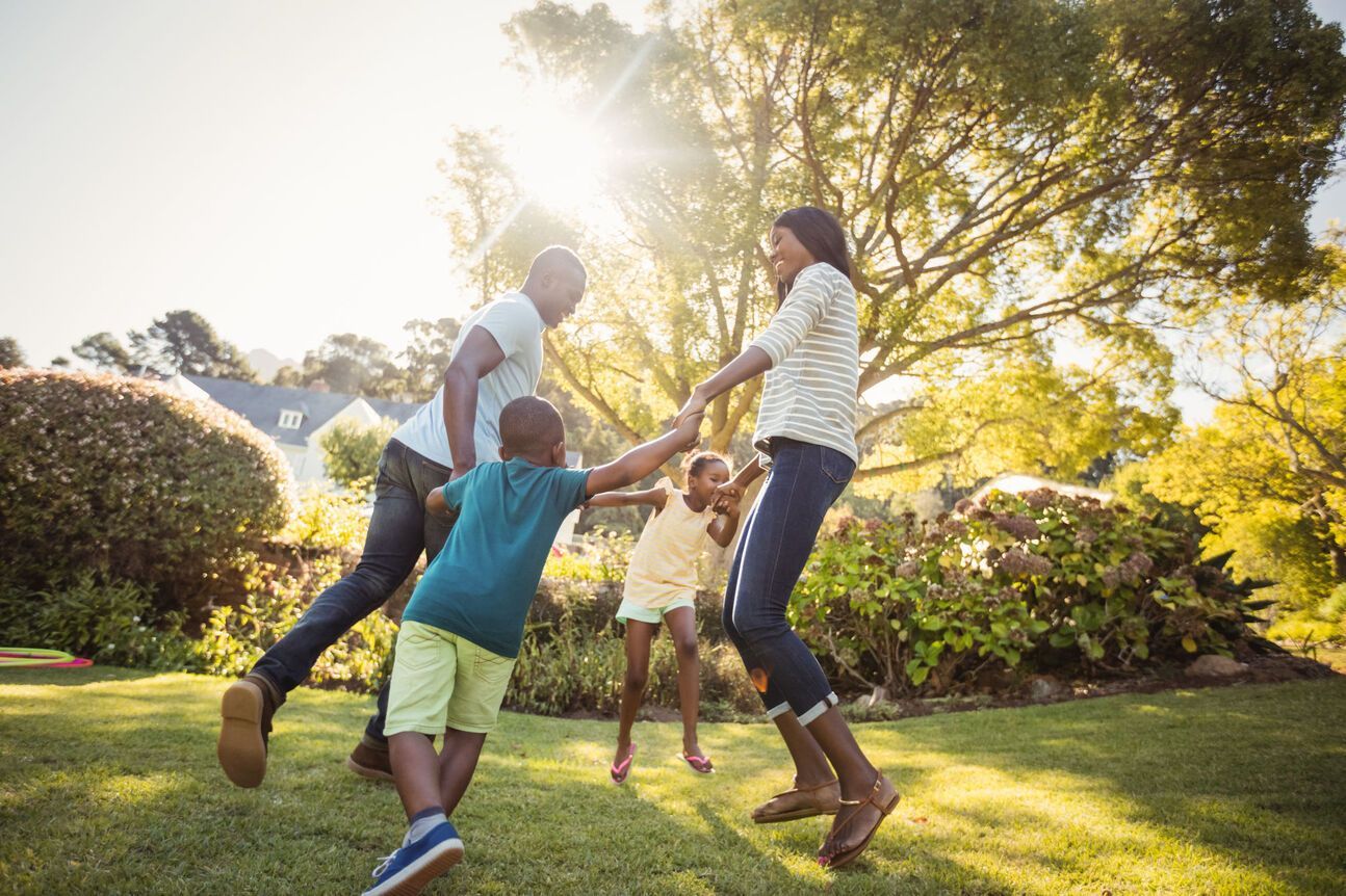 A Black family of four playing on the grass in a sunny yard, holding hands and spinning.