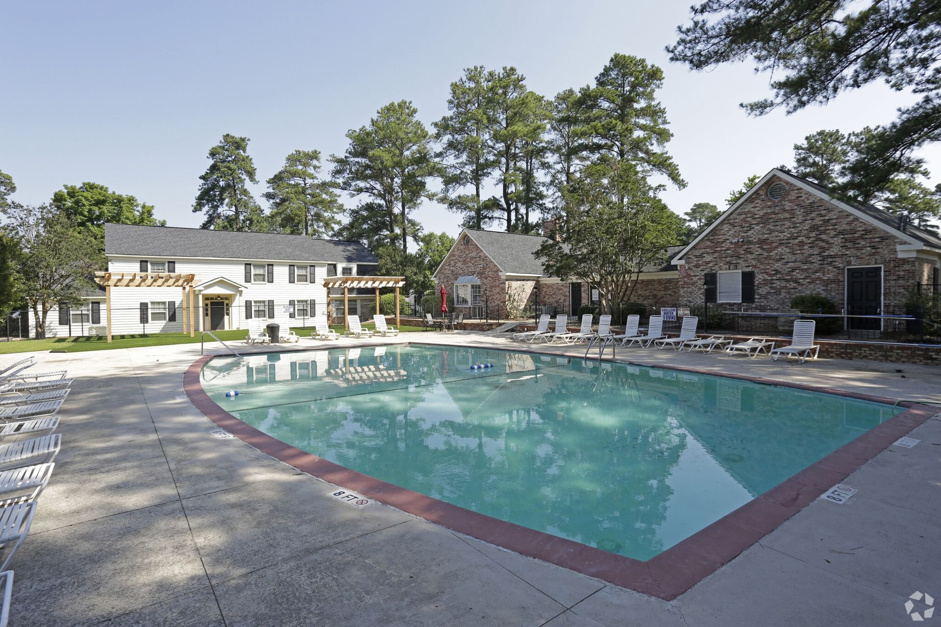 Swimming pool with lounge chairs and buildings in the background under a sunny sky.