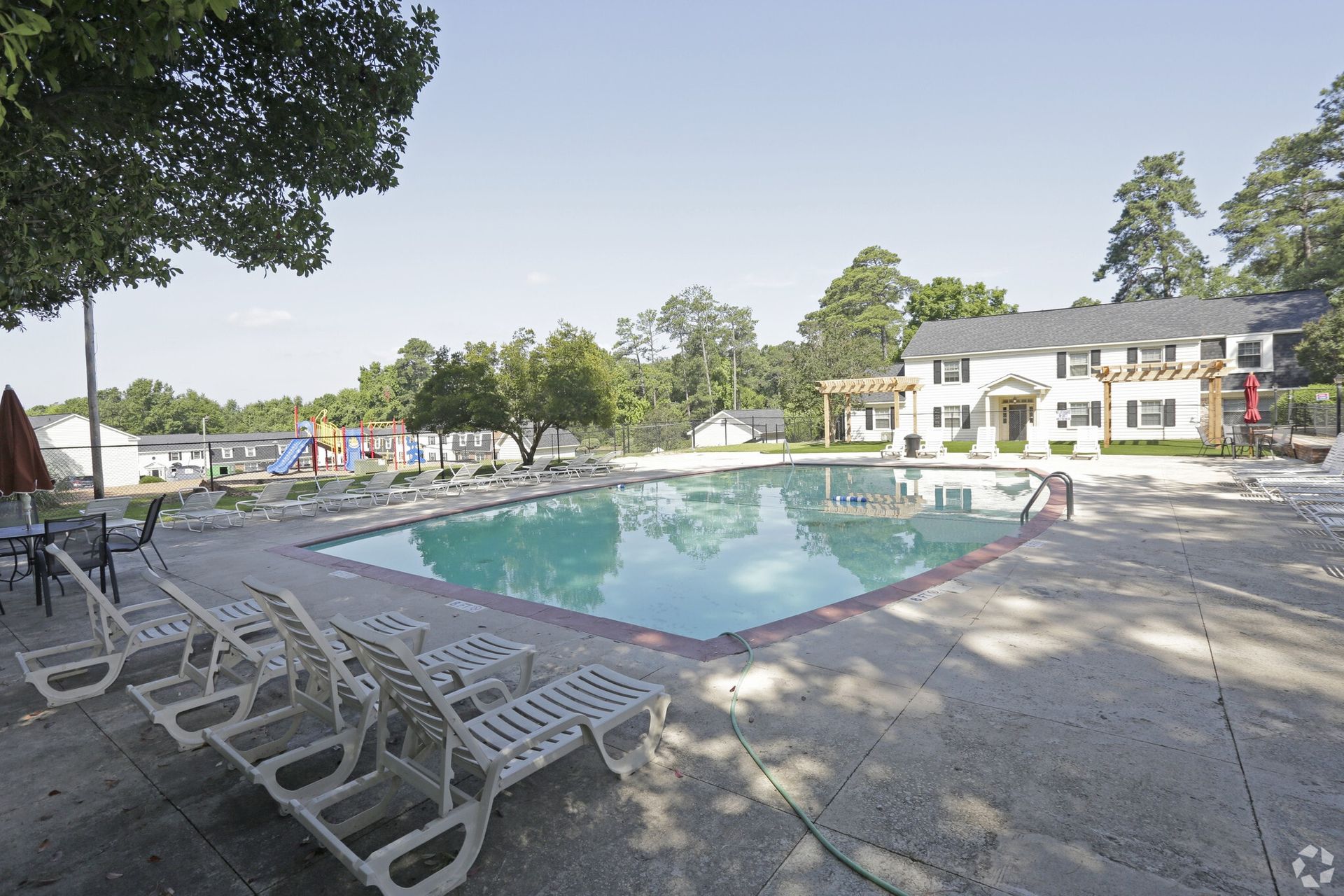 Pool with lounge chairs, trees, and a building in the background on a sunny day.