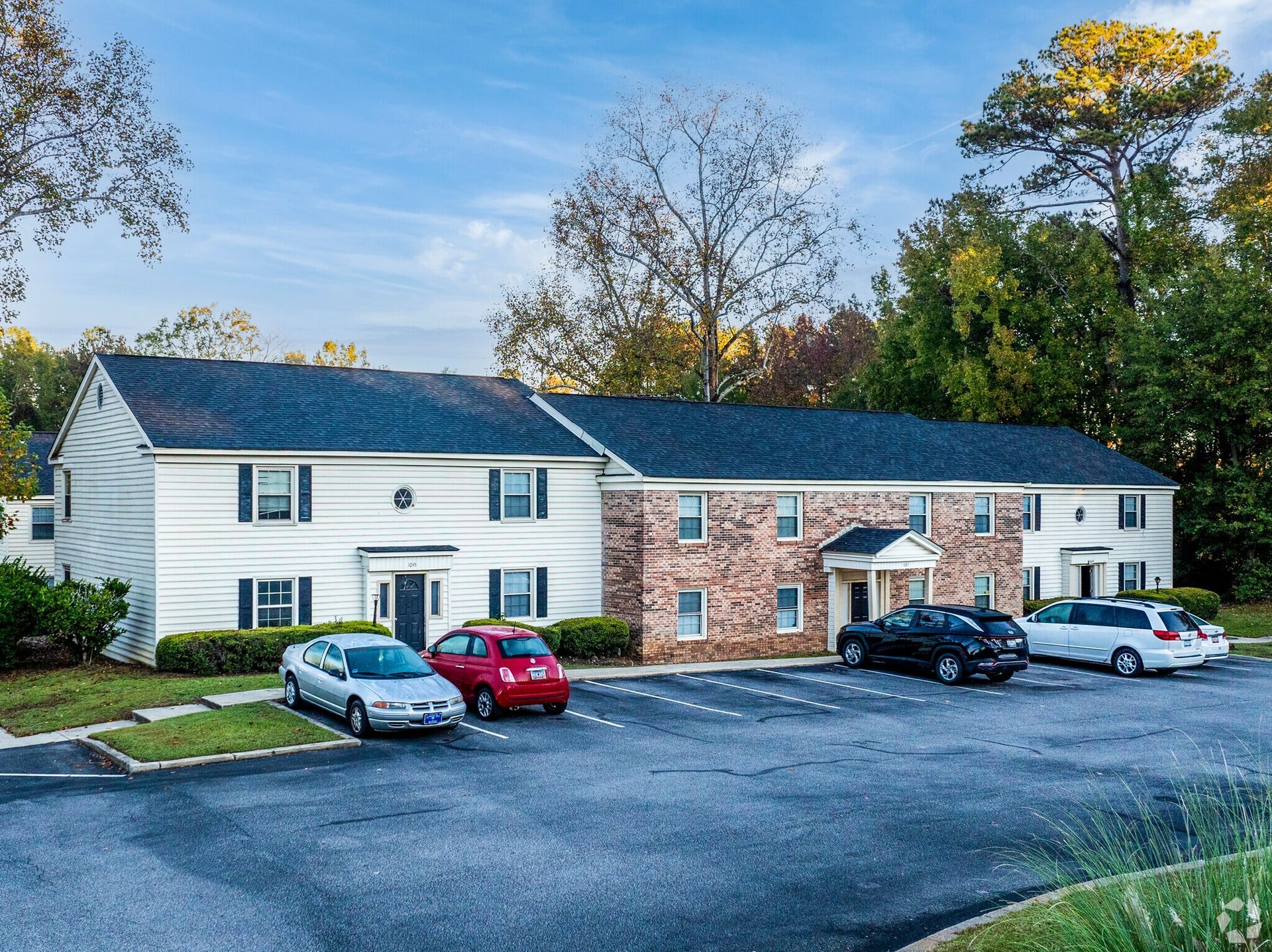 Apartment complex with brick and white siding. Cars parked in front. Blue sky and trees in background.