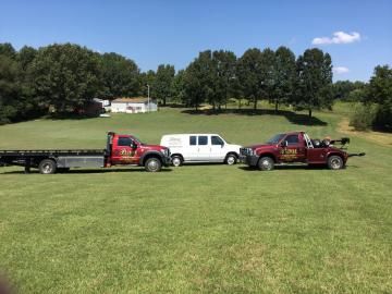 Two tow trucks and a white van on a grassy field in front of trees and a house on a sunny day.