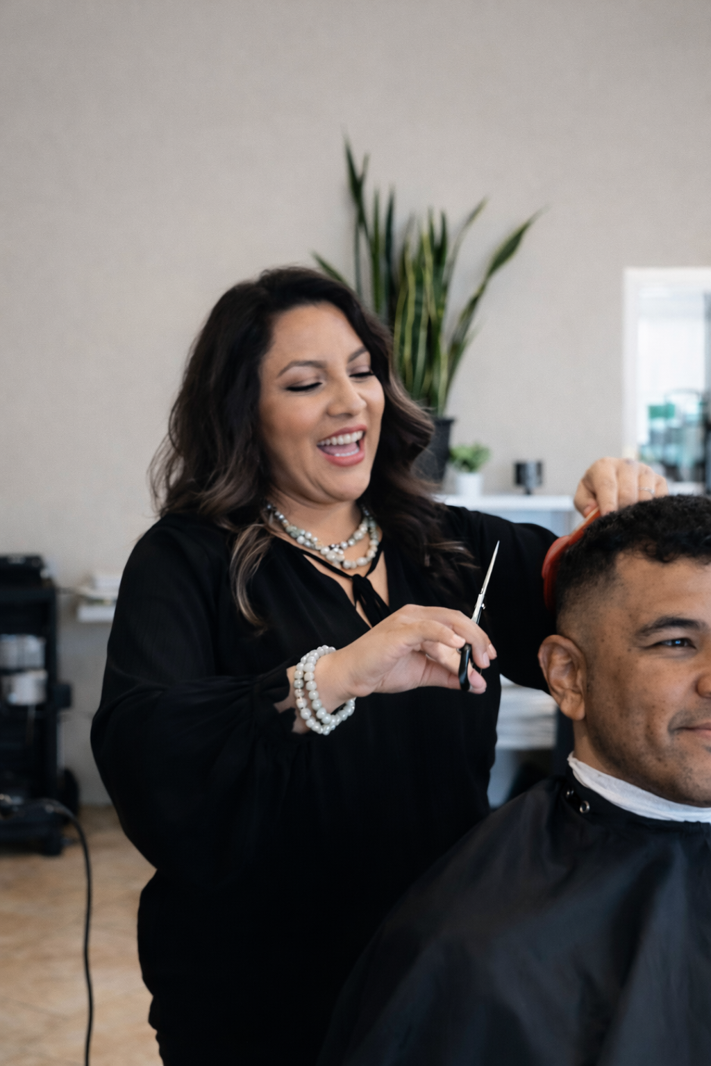 Hairdresser cuts a client's hair in a salon. The hairdresser smiles, wearing black. The client wears a cape.
