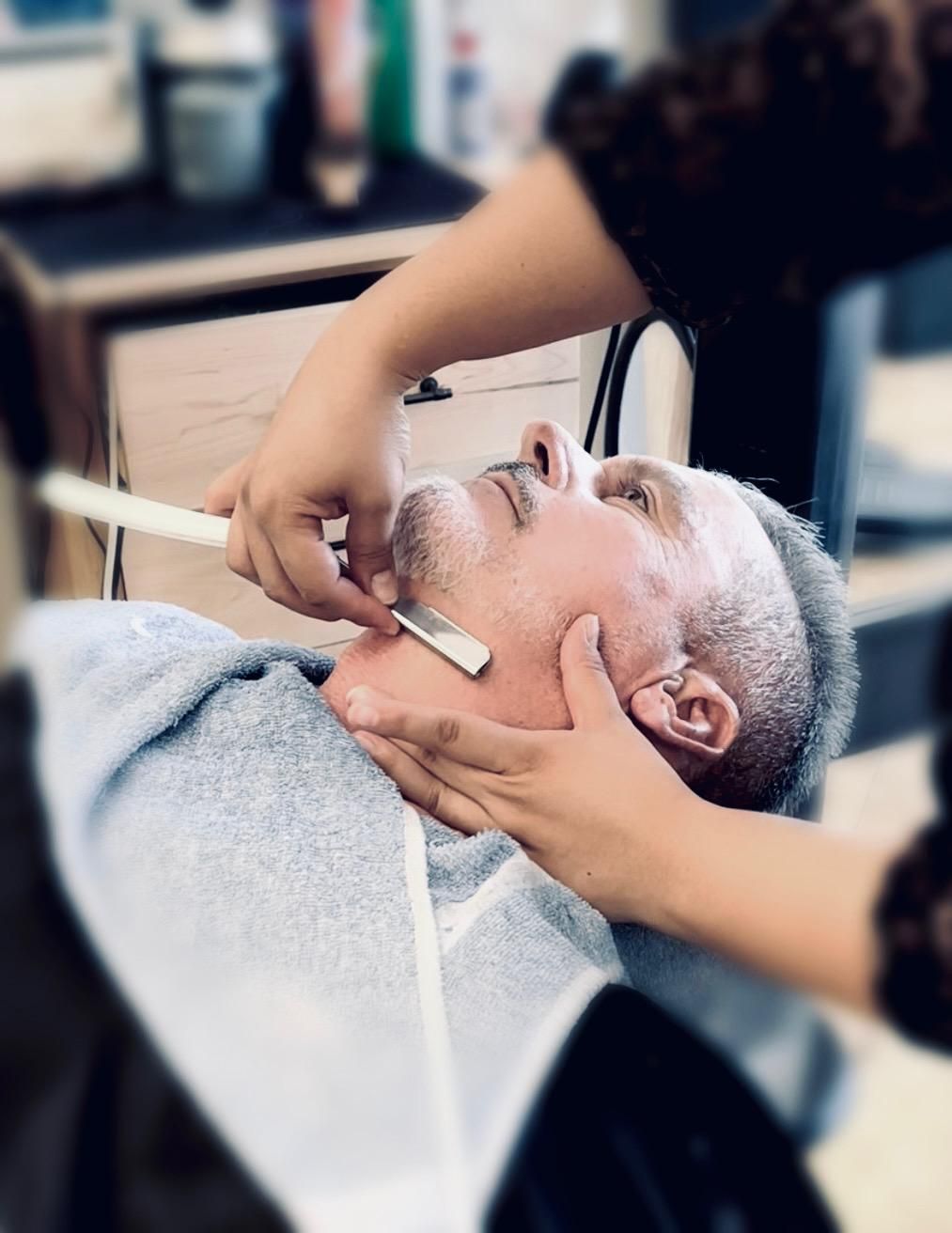 Person getting a straight razor shave at a barbershop. Razor held to jaw, face tilted up.