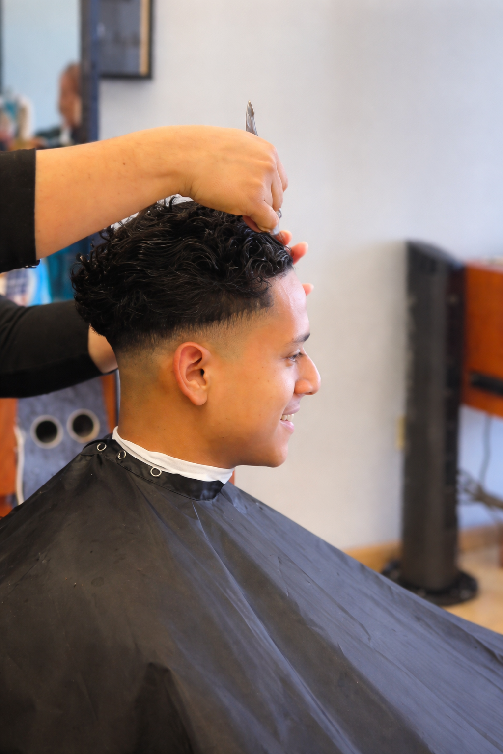 Man getting a haircut at a barbershop; black cape, faded side and top with curly hair. Smiling.