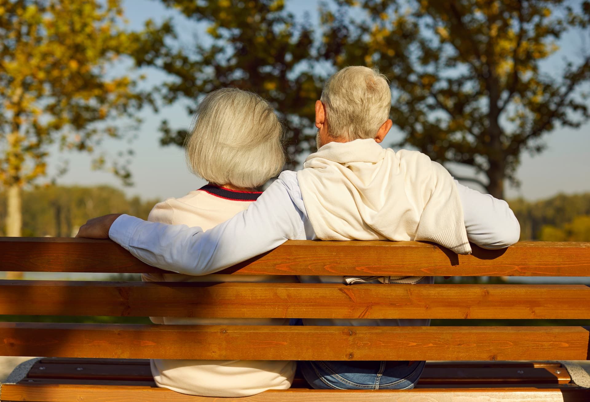 Two people sit side-by-side on a wooden bench outdoors, arms around each other, facing a background of trees.