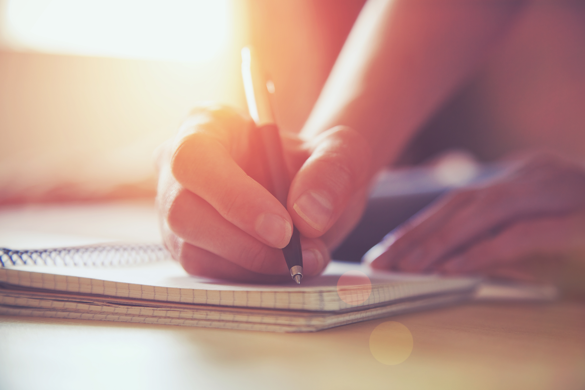 Close-up of a person’s hand writing in a spiral notebook, illuminated by warm, soft sunlight.