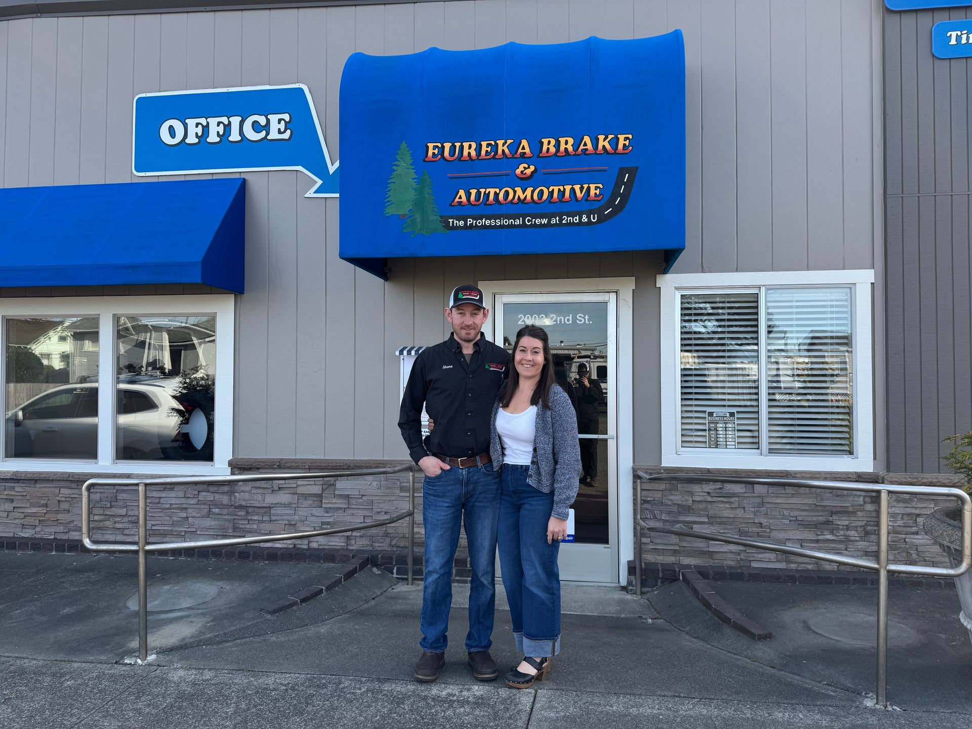 A person in a black shirt and a person in a white top stand together in front of the Eureka Brake & Automotive office