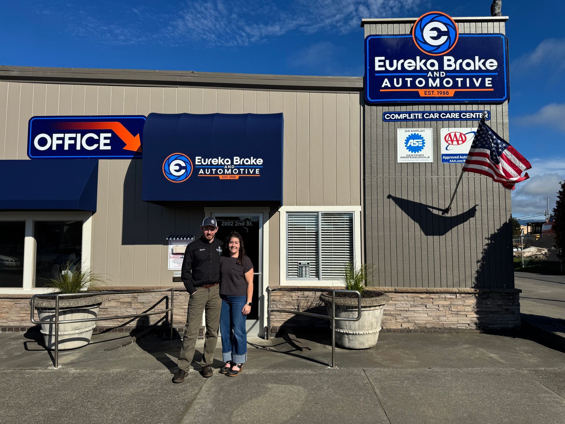A man and a woman stand outside Eureka Brake and Automotive under a blue awning with a small American flag on the wall | Eureka Brake and Automotive