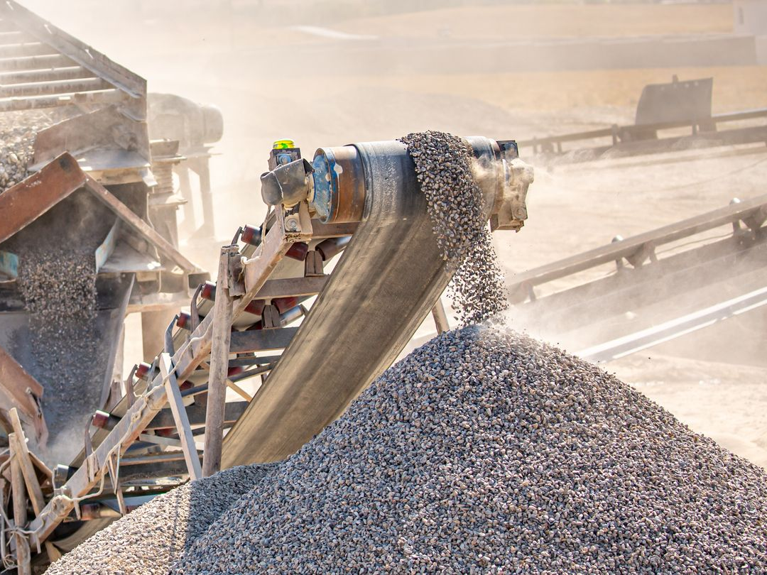 Industrial conveyor dumping gray gravel into a large pile in a dusty quarry setting