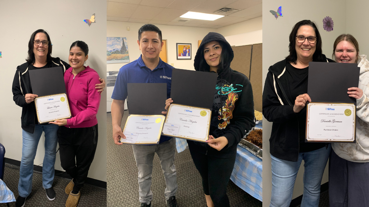 Three people in an office hold certificates and flowers, smiling during a recognition celebration.