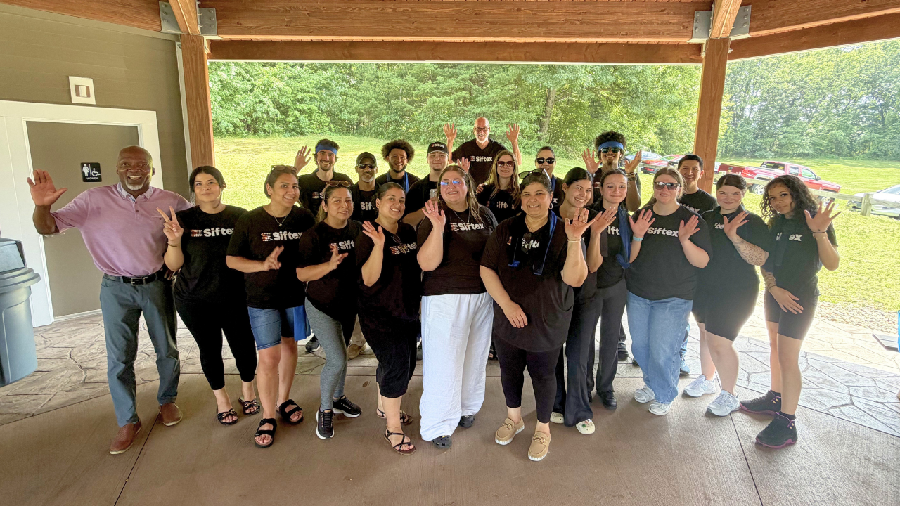 Group of people in black shirts posing under a pavilion, smiling and holding up hand signs.