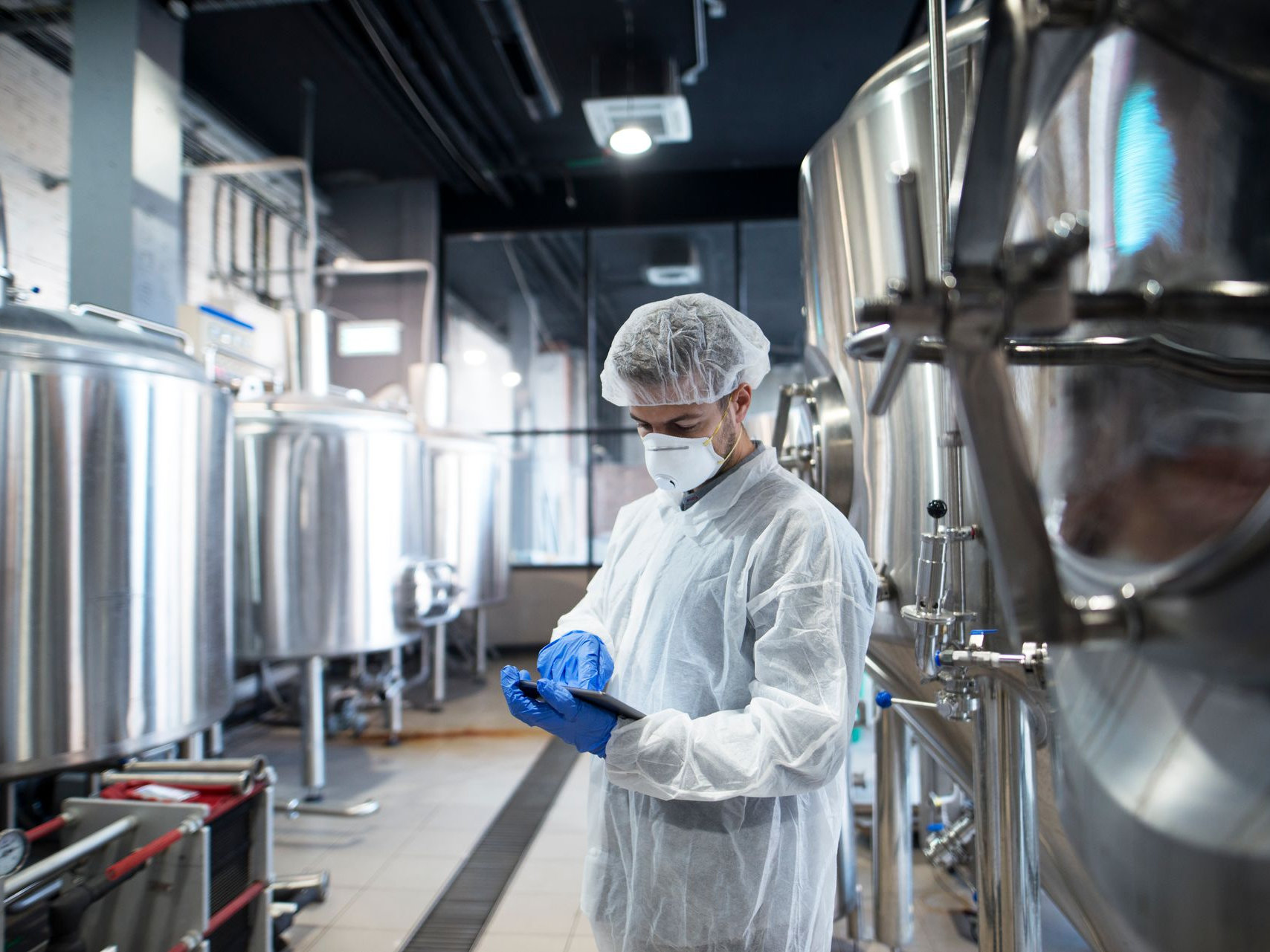 Lab technician in white protective gear and blue gloves inspecting equipment in a stainless steel facility