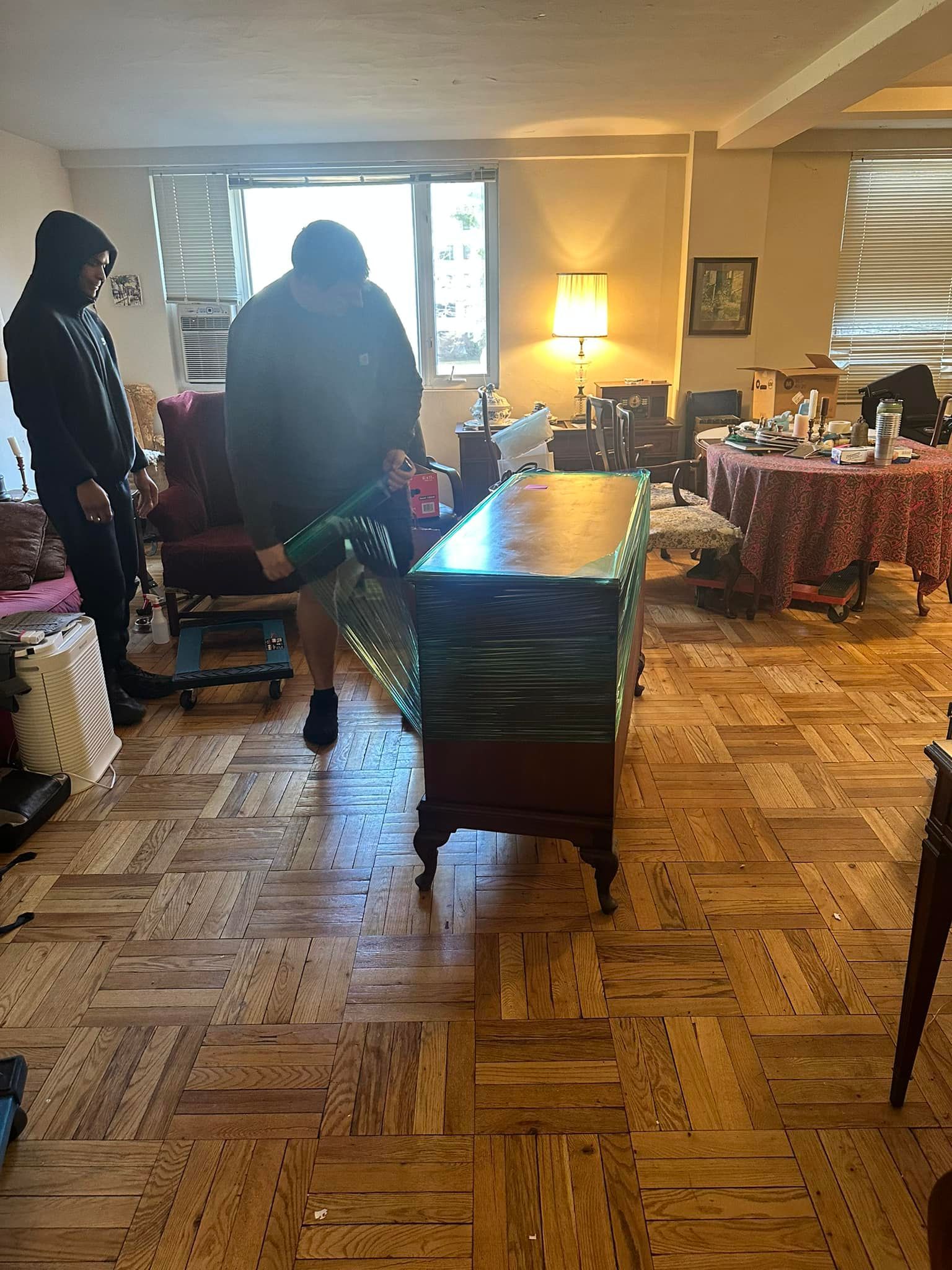 Two people moving furniture, wrapping a wood cabinet in plastic, indoor setting.