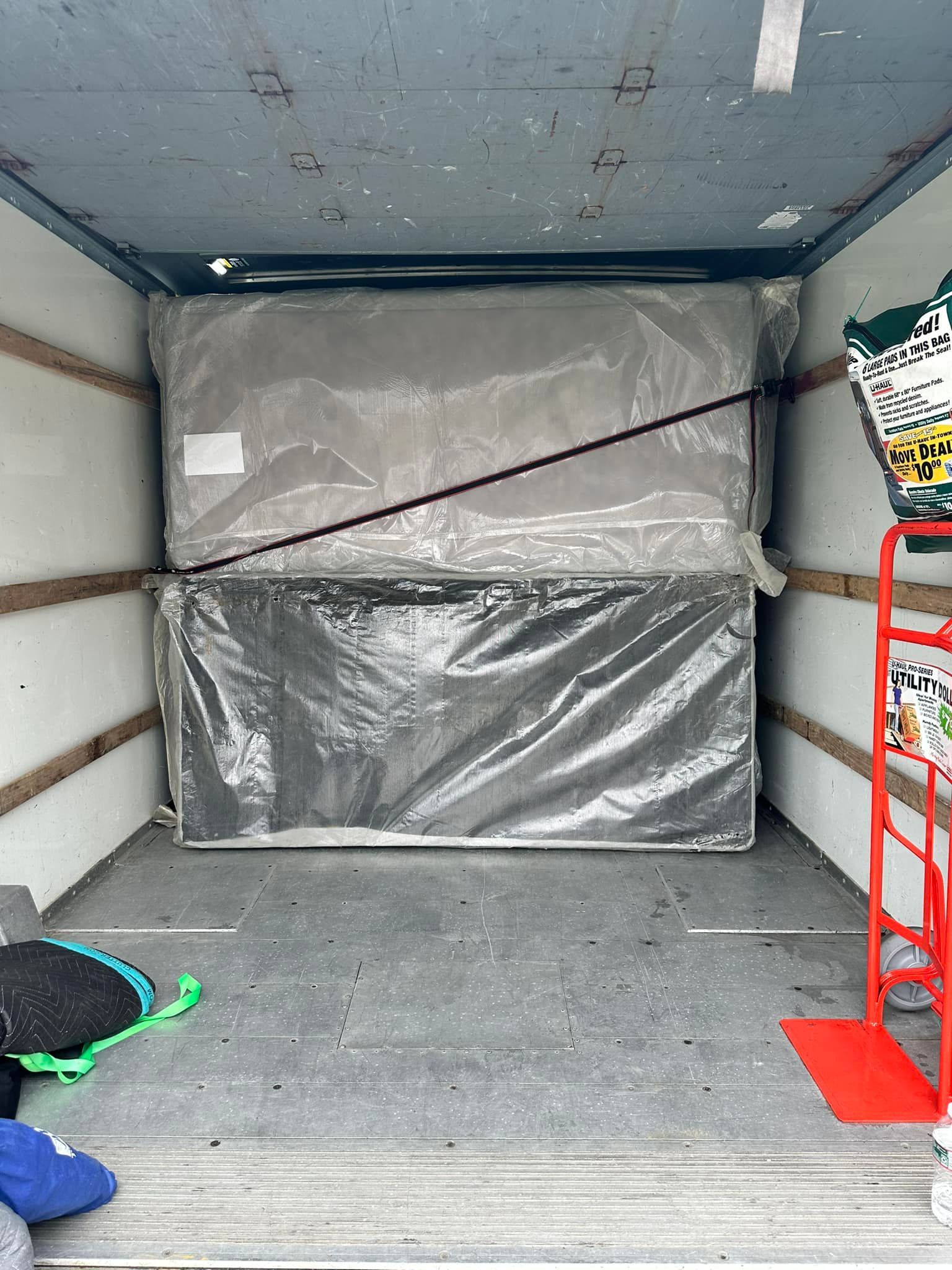 Mattresses wrapped in plastic inside a moving truck, secured with a black strap.