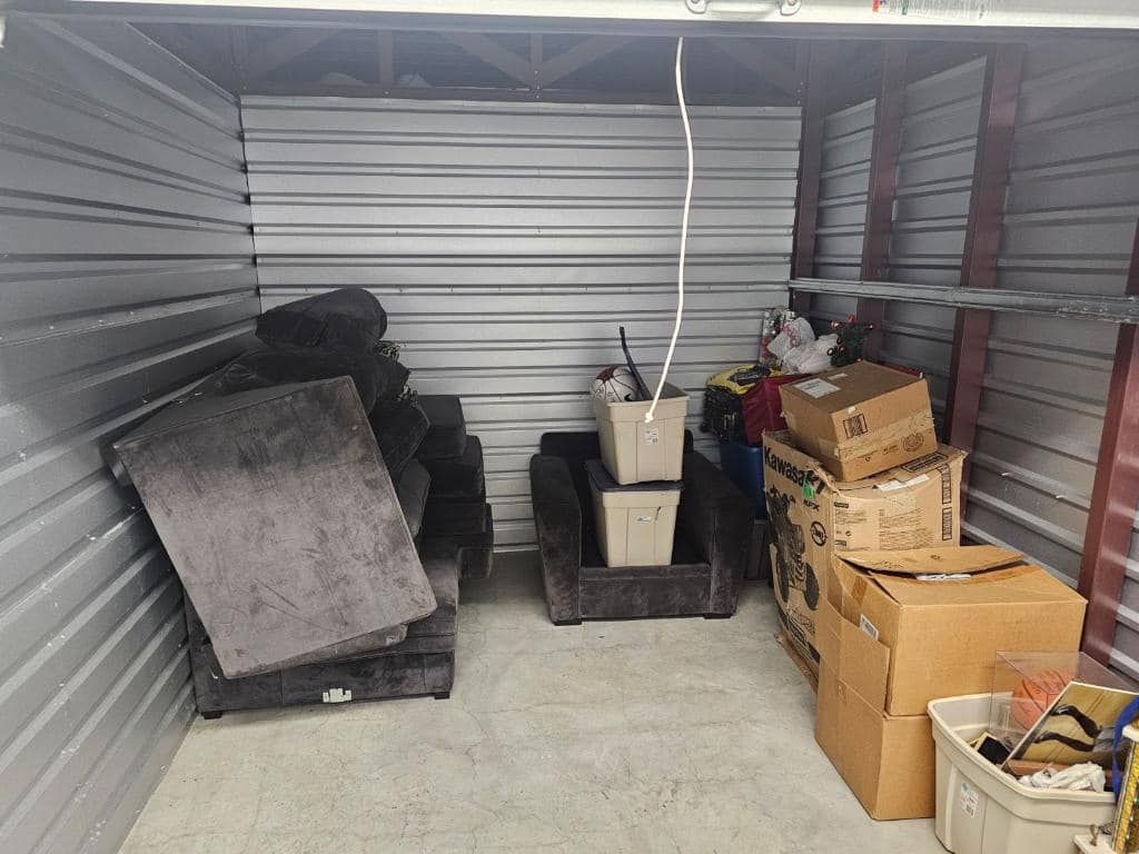 A storage unit filled with gray furniture, boxes, and miscellaneous items.