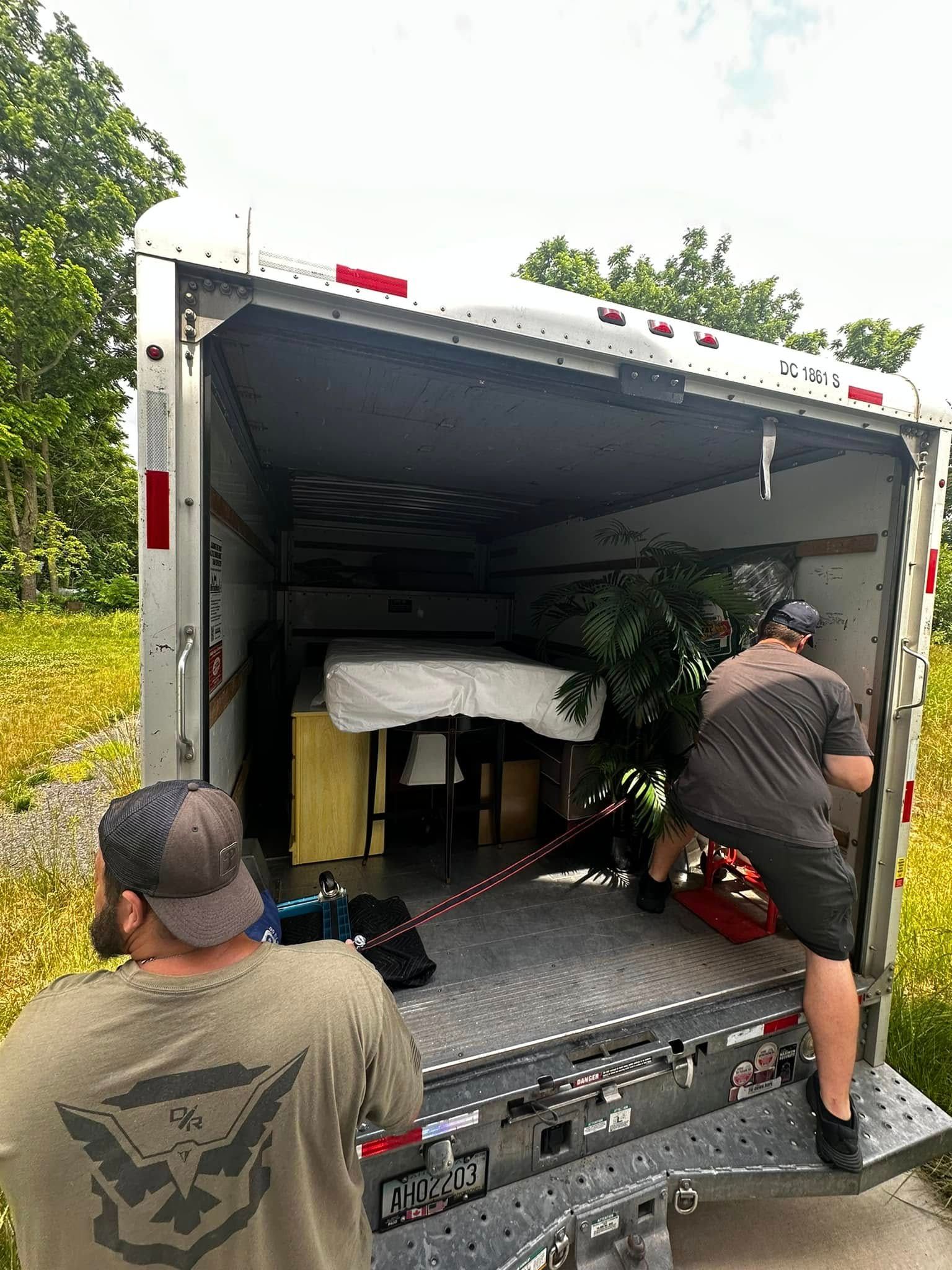 Two men loading a truck: furniture, a plant, and a mattress. Outdoors, near a grassy area.