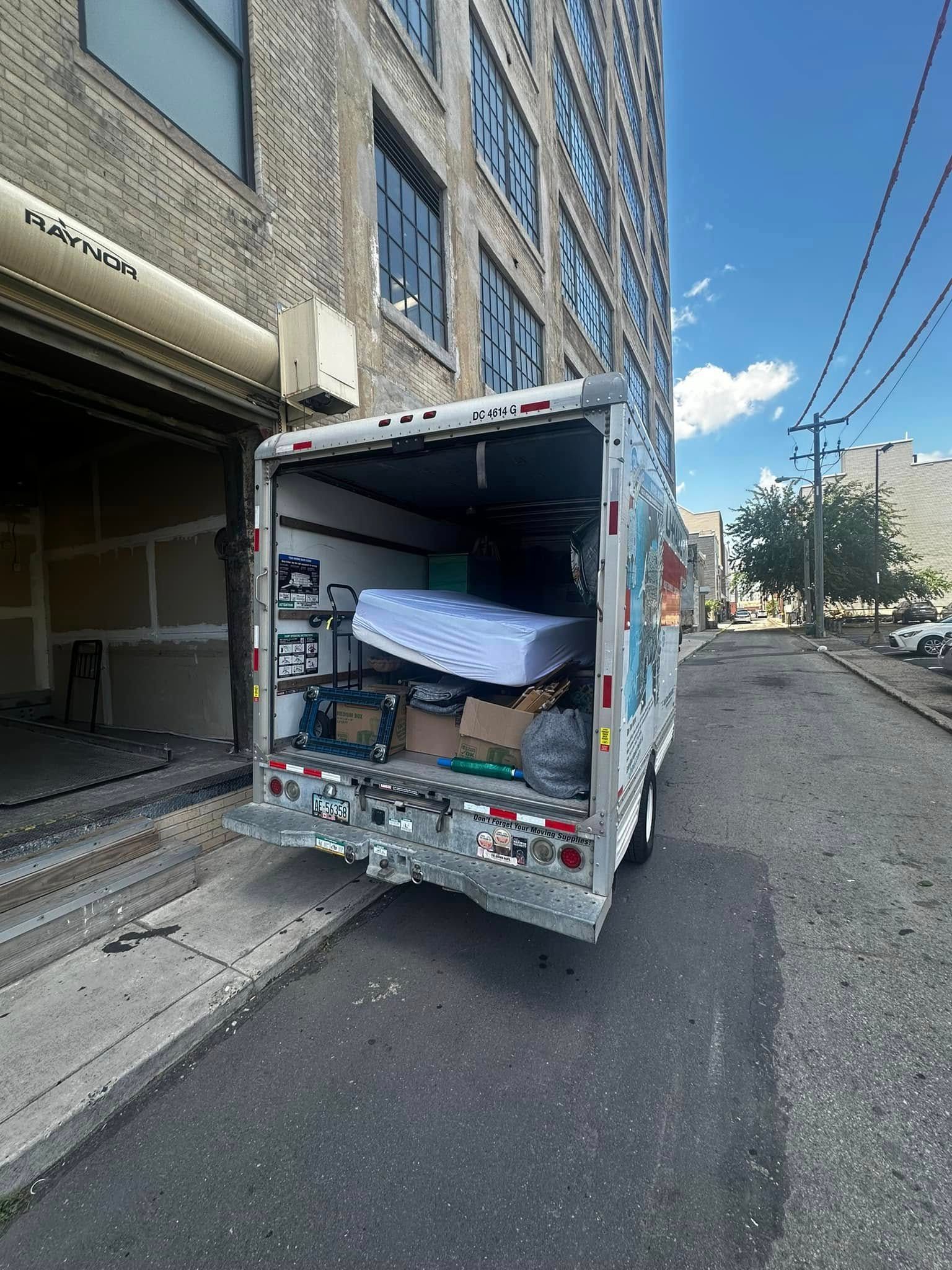 Moving truck backed into an alley, open, loaded with a mattress and other items, near a brick building.