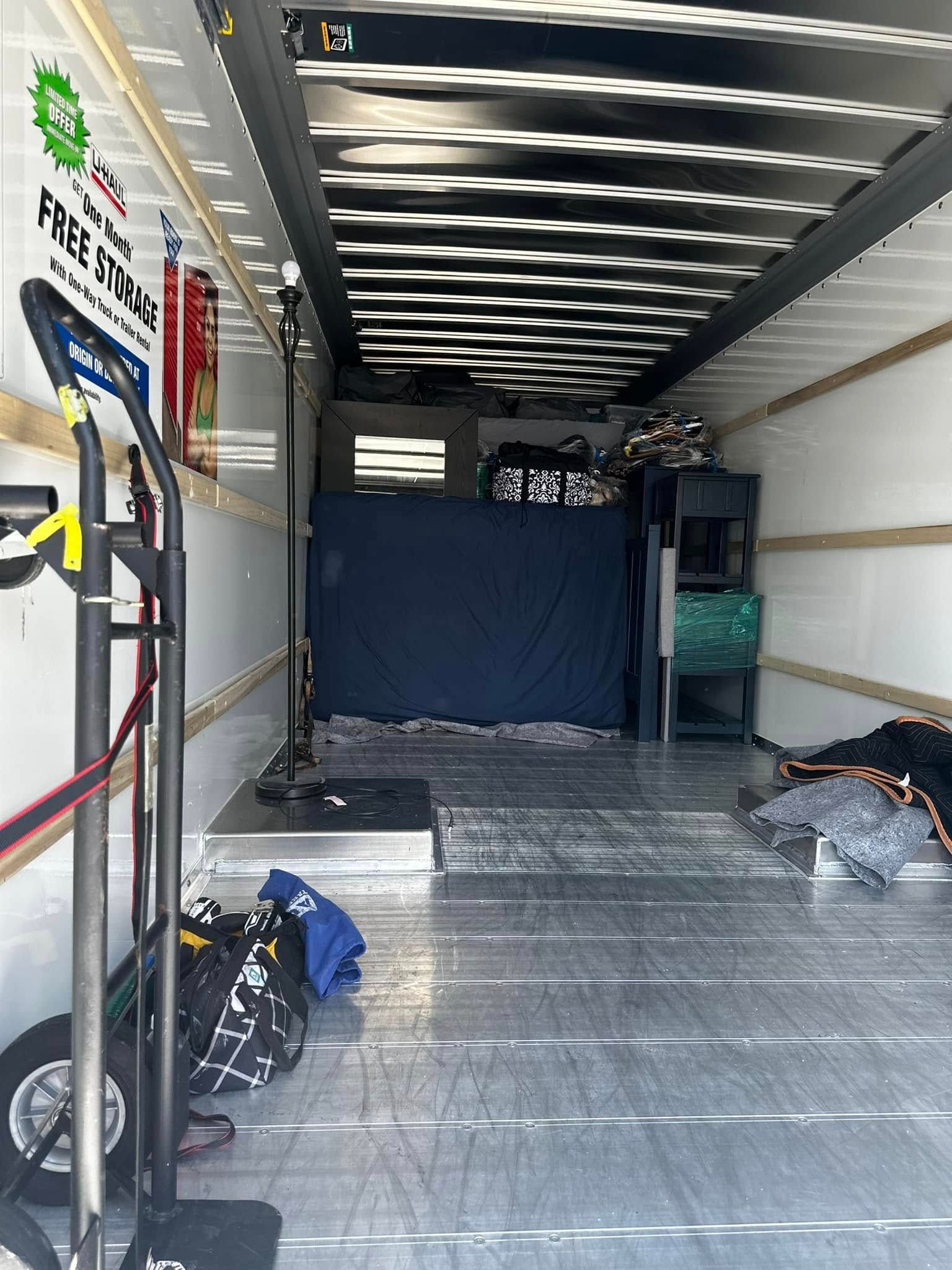 Interior of a moving truck, mostly empty. A hand truck, a mattress, and boxes are visible.