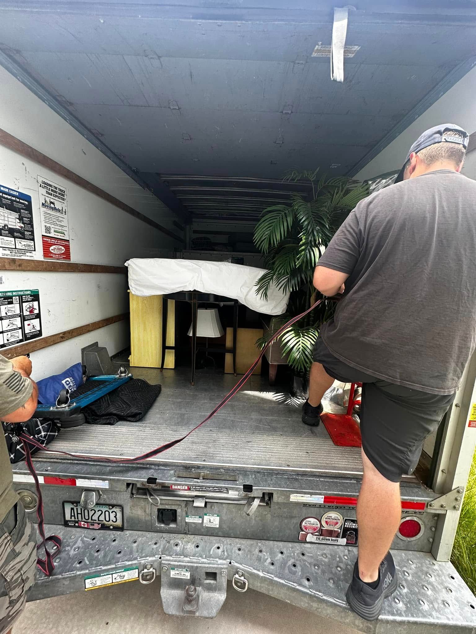People loading furniture into a moving truck, including a bed and a large plant.