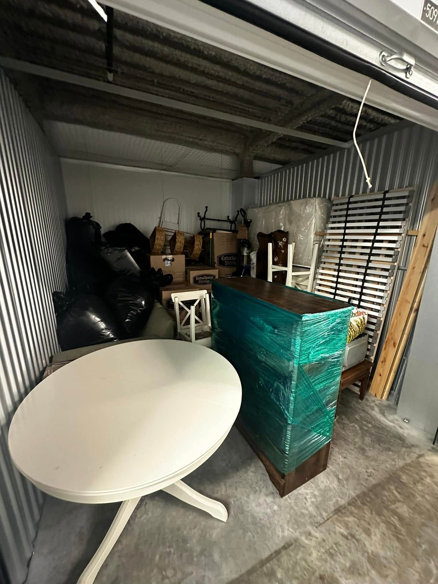 Storage unit with a white table, boxes, furniture, and black bags.