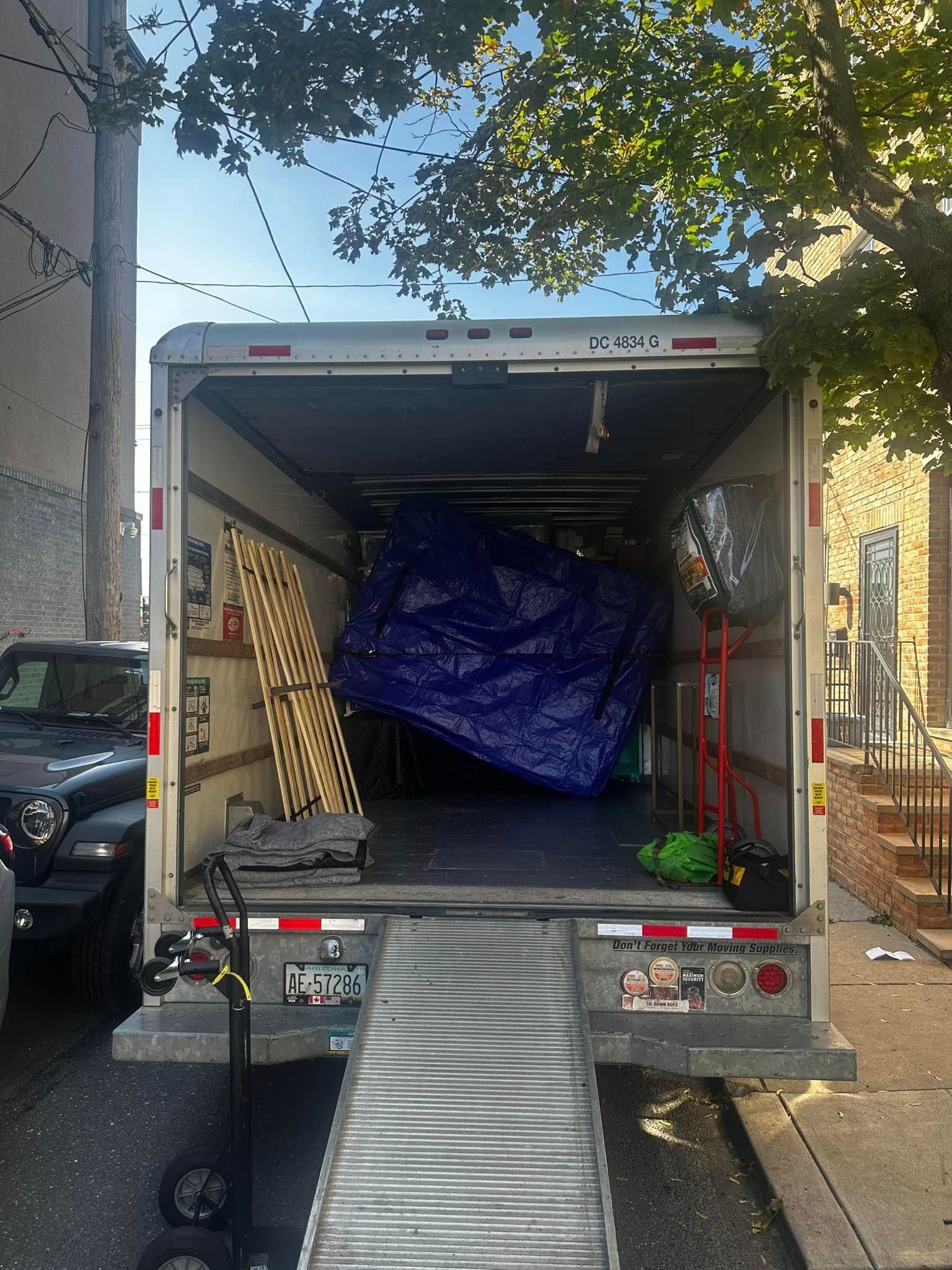 Truck bed open, loading ramp down, furniture covered in blue tarp, wood planks, and other items visible.