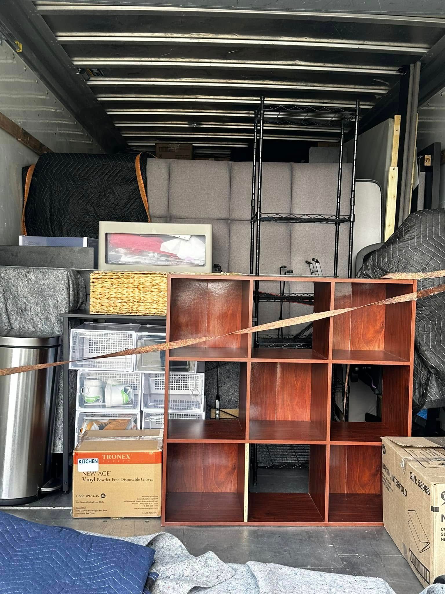 Interior of a moving truck packed with furniture, boxes, and appliances, including a brown cube shelf.