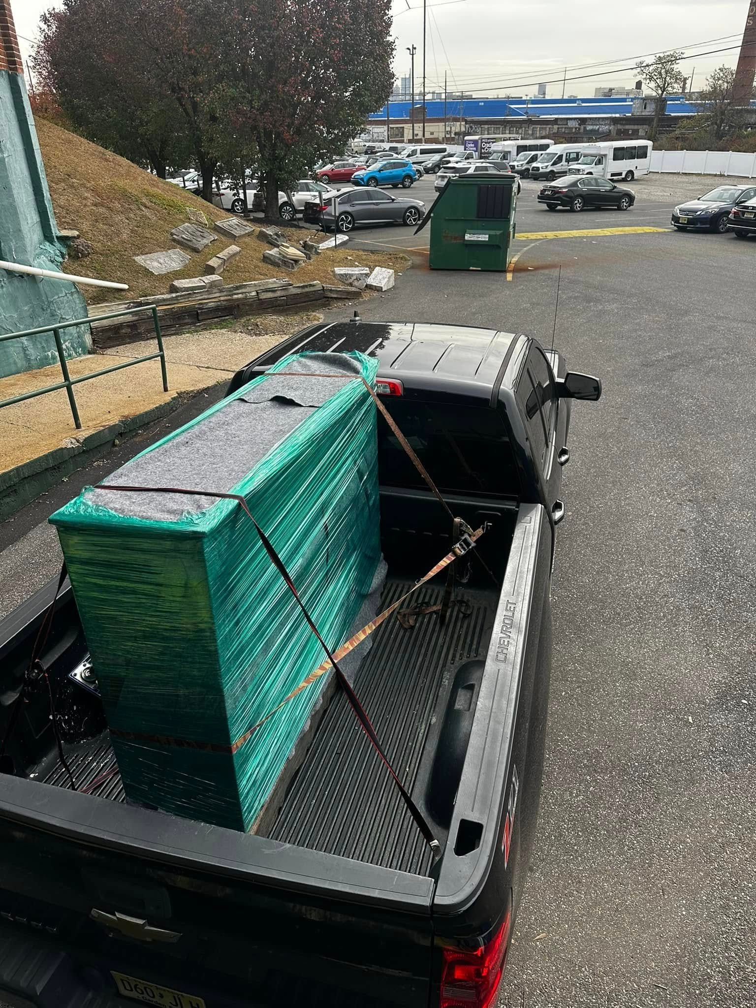 Black pickup truck bed carrying a tall, turquoise-green box. Dumpster and cars in the background.