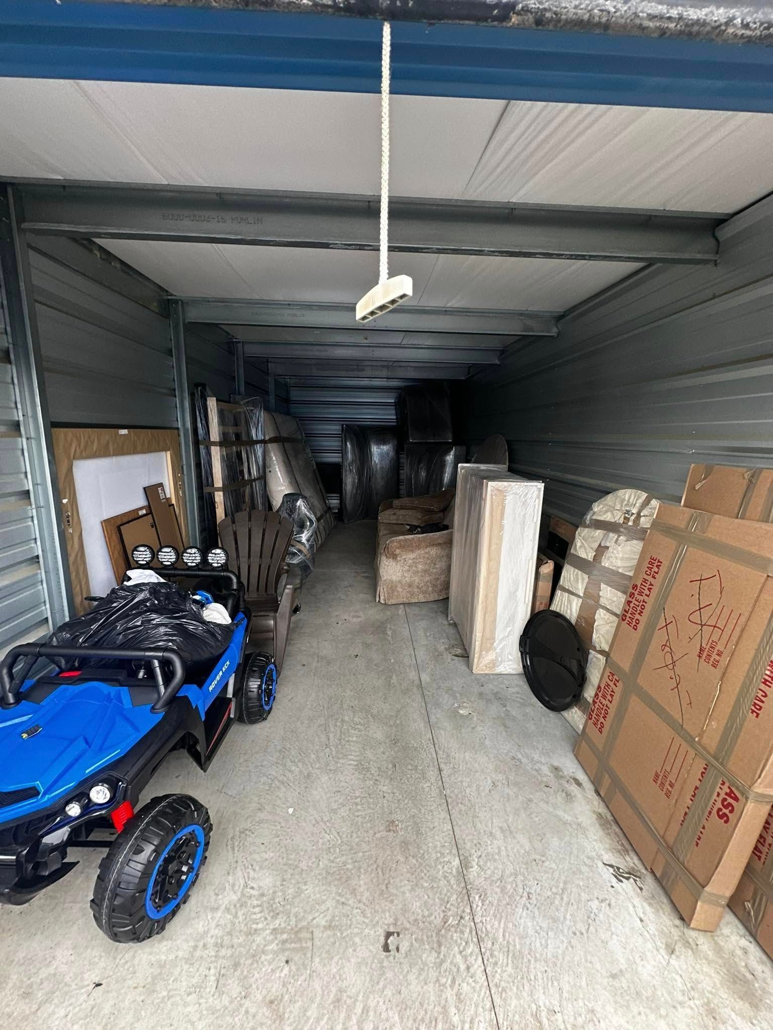 Inside a packed storage unit; a blue toy car, boxes, and wrapped furniture line the space.