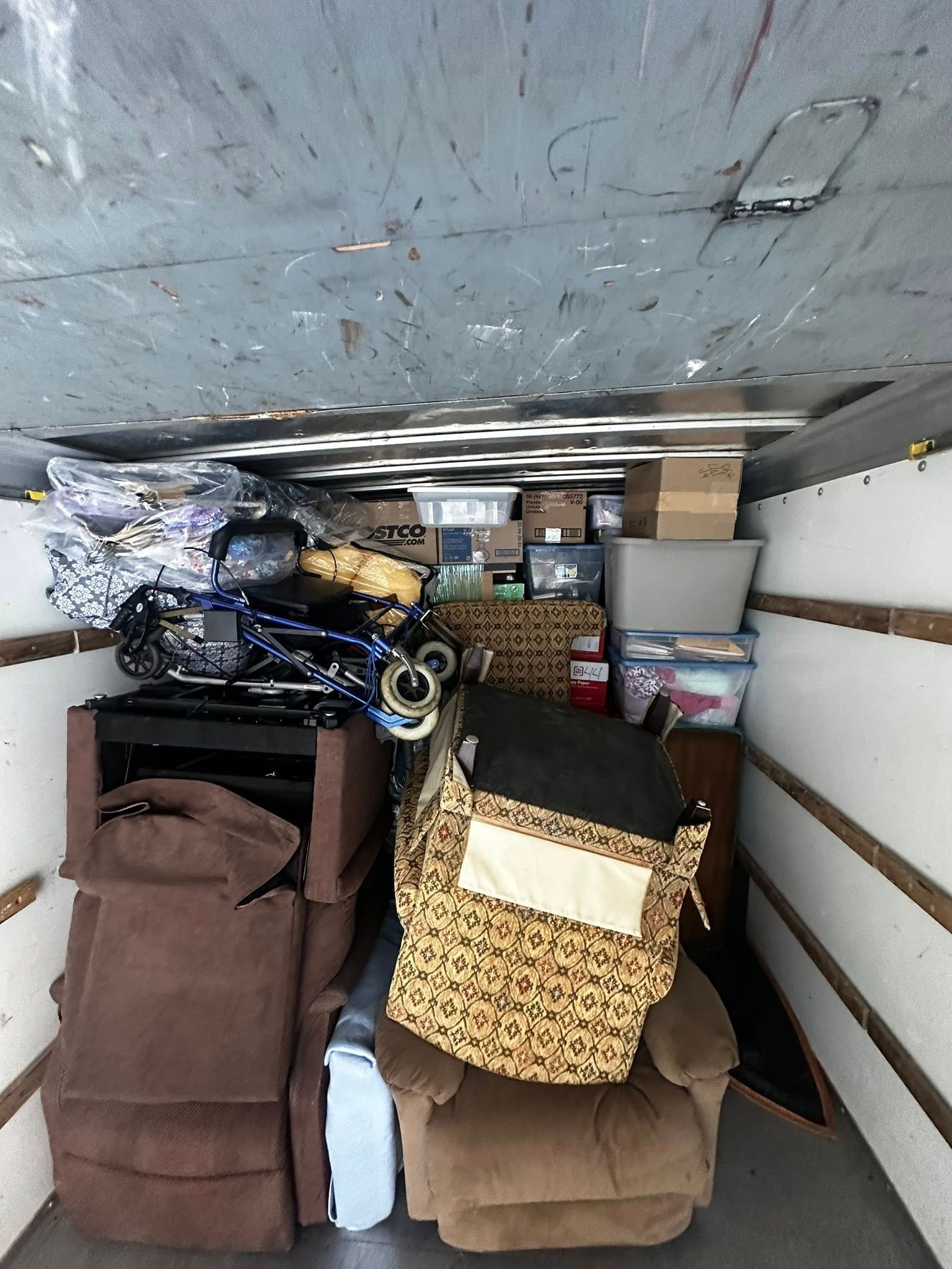 Moving truck interior packed with furniture, boxes, and belongings; brown recliner, leopard-print chair visible.