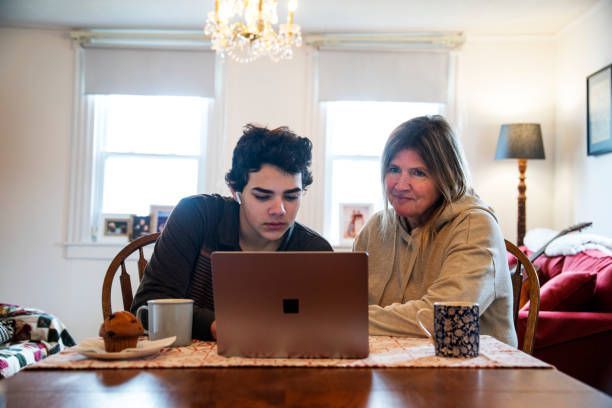 Teacher assisting a student in a classroom. The teacher is pointing to the student's notebook. Other students are in the background.