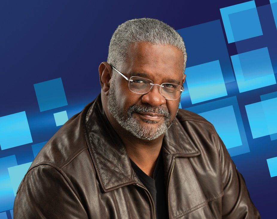 Man with glasses and grey beard in leather jacket, smiling against a blue geometric background.