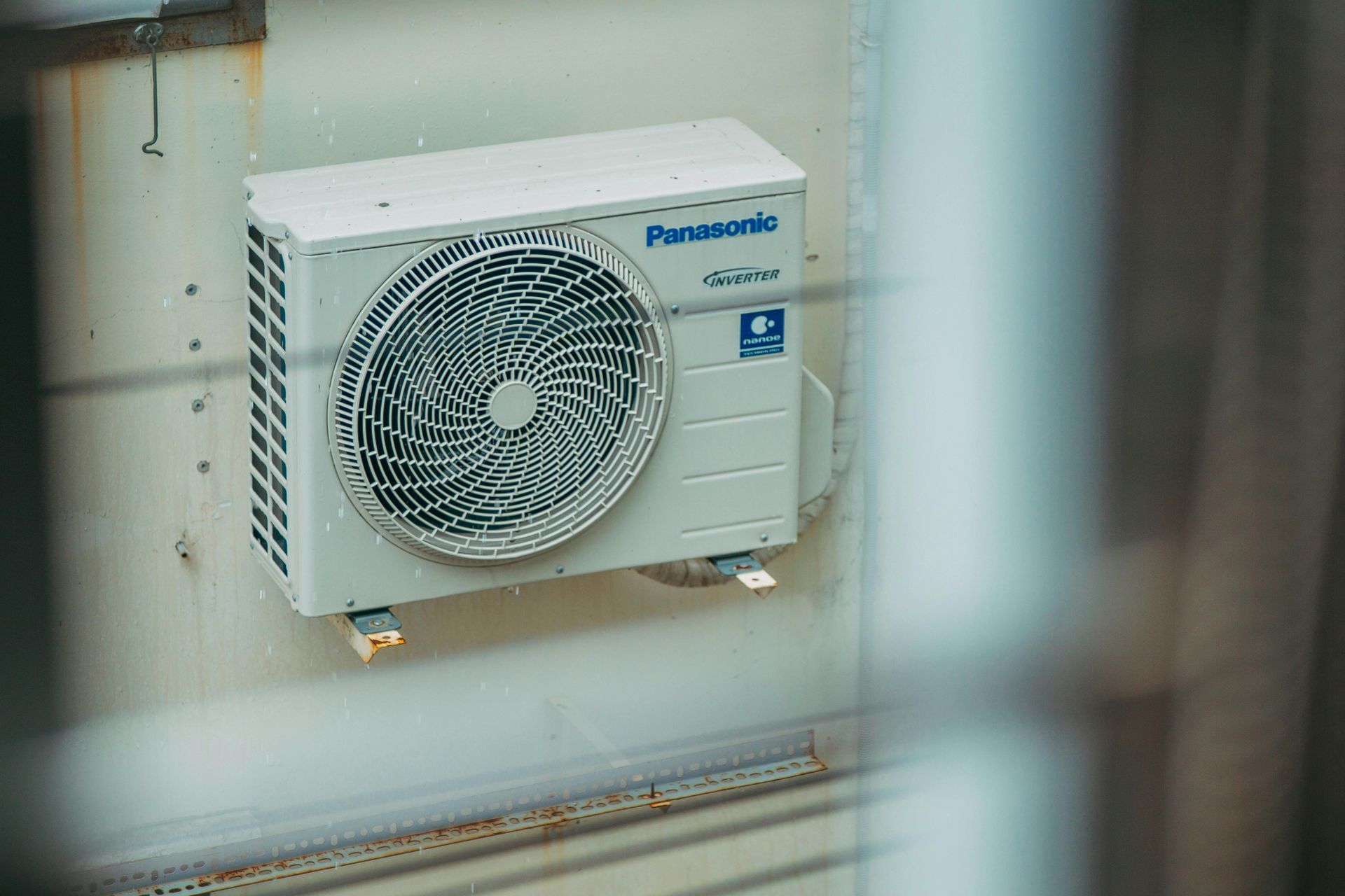 White Panasonic outdoor air conditioner unit mounted on a wall behind a window.