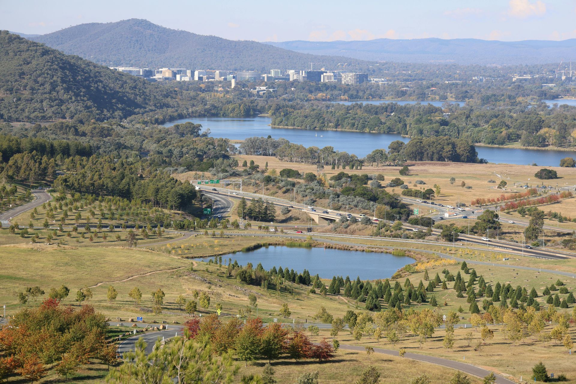 High-angle landscape view of a scenic park with a pond, lush trees, rolling hills, and a lake under a clear blue sky.