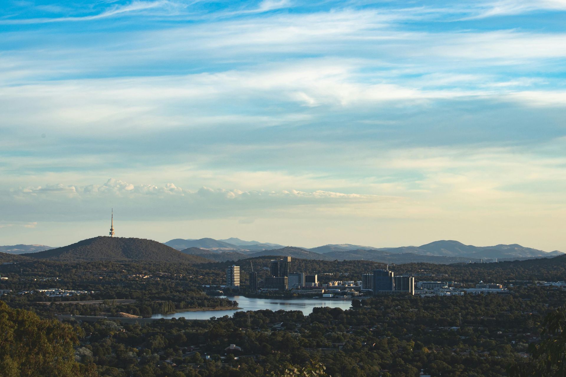 A cityscape of Canberra, Australia, featuring a lake and Black Mountain Tower under a soft, cloudy sky.