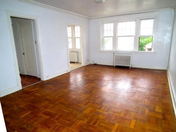 A bright, empty living room with hardwood floors and white walls; a doorway and windows are visible.