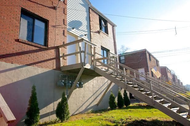 Red brick apartment building with wooden stairs and deck leading to a second-floor entrance, green grass.
