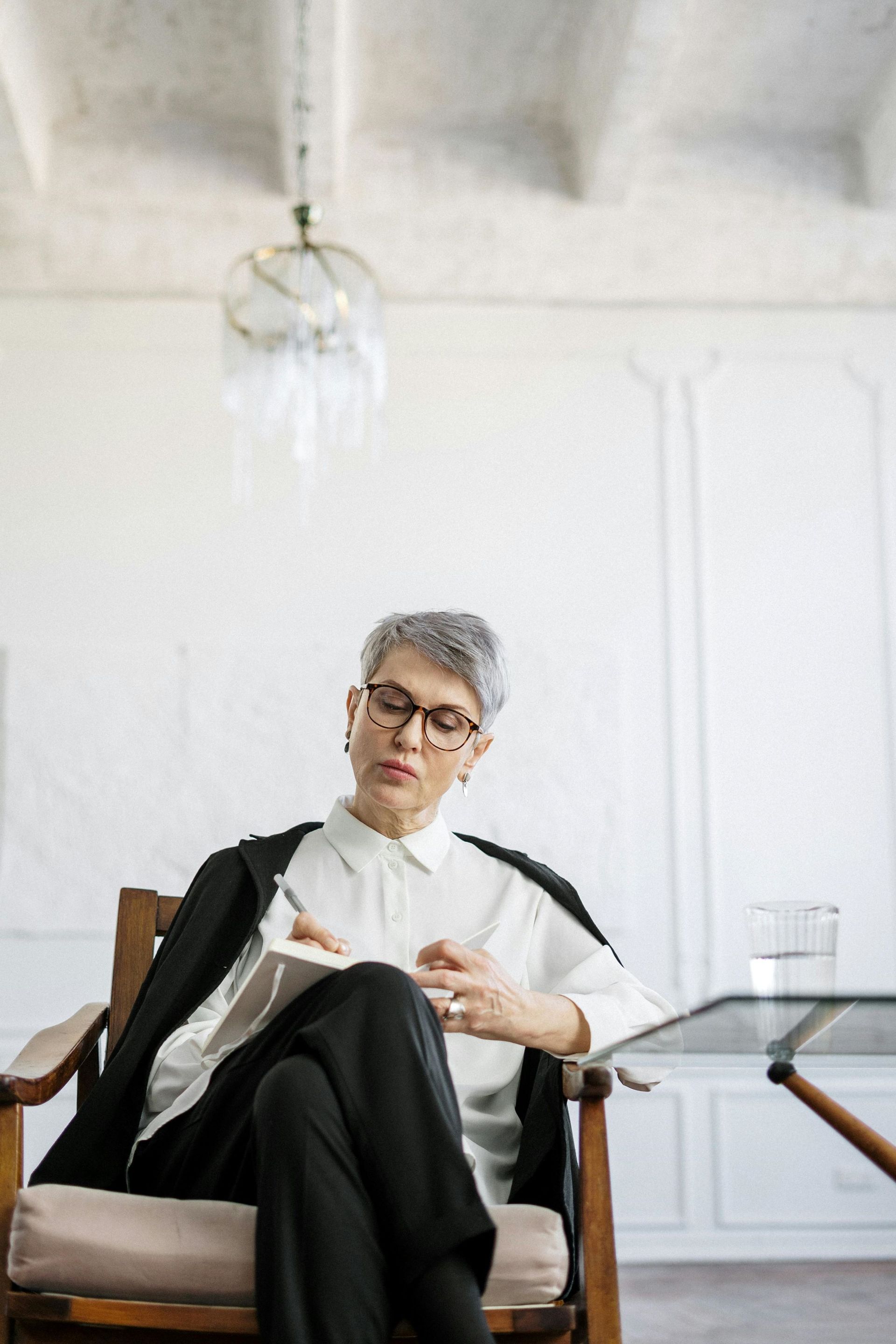 A woman in white suit and wearing glasses is sitting with legs crossed in front of her while writing