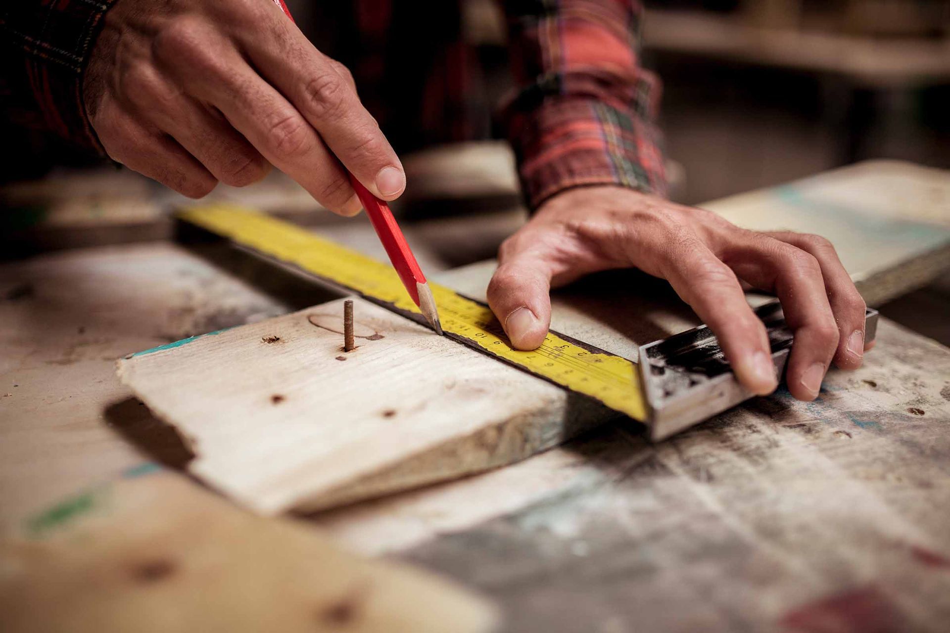 Hands measuring and marking a wooden board with a yellow ruler and red pencil in a workshop