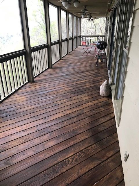 Covered wooden porch with railings, a chair, and a bag along the wall, viewed down a long screened walkway