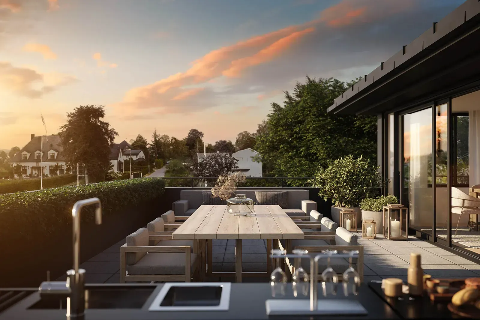Romslig takterrasse med lang spisebord og utekjøkken, omgitt av hekk og utsikt over et etablert boligområde ved solnedgang