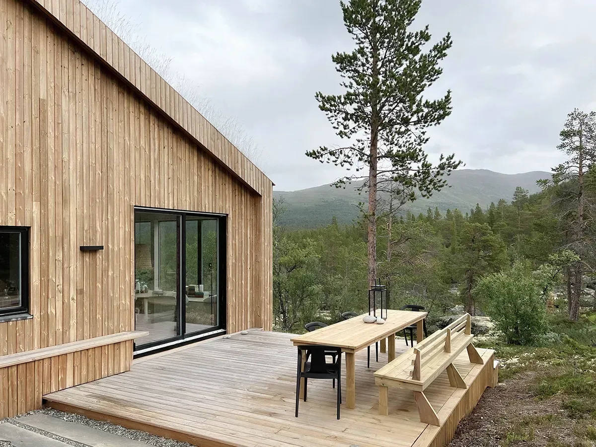 Terrasse med plassbygget benk og spisebord i tre, vendt mot utsikten til fjellskog og dalsøkk.