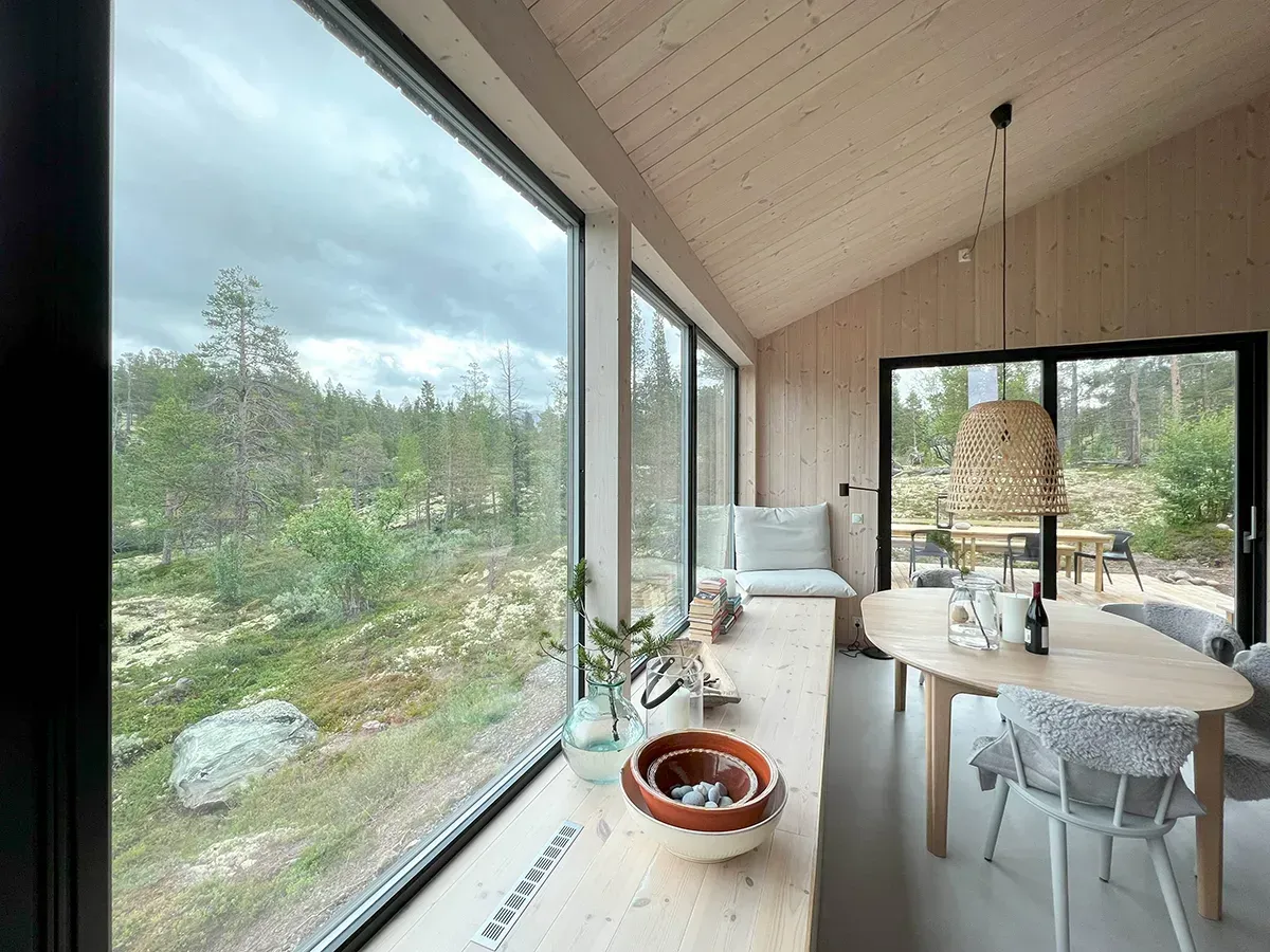 Interiør med store vinduer og gjennomgående bruk av lys trekledning i tak og vegger, med direkte kontakt til omgivende natur.