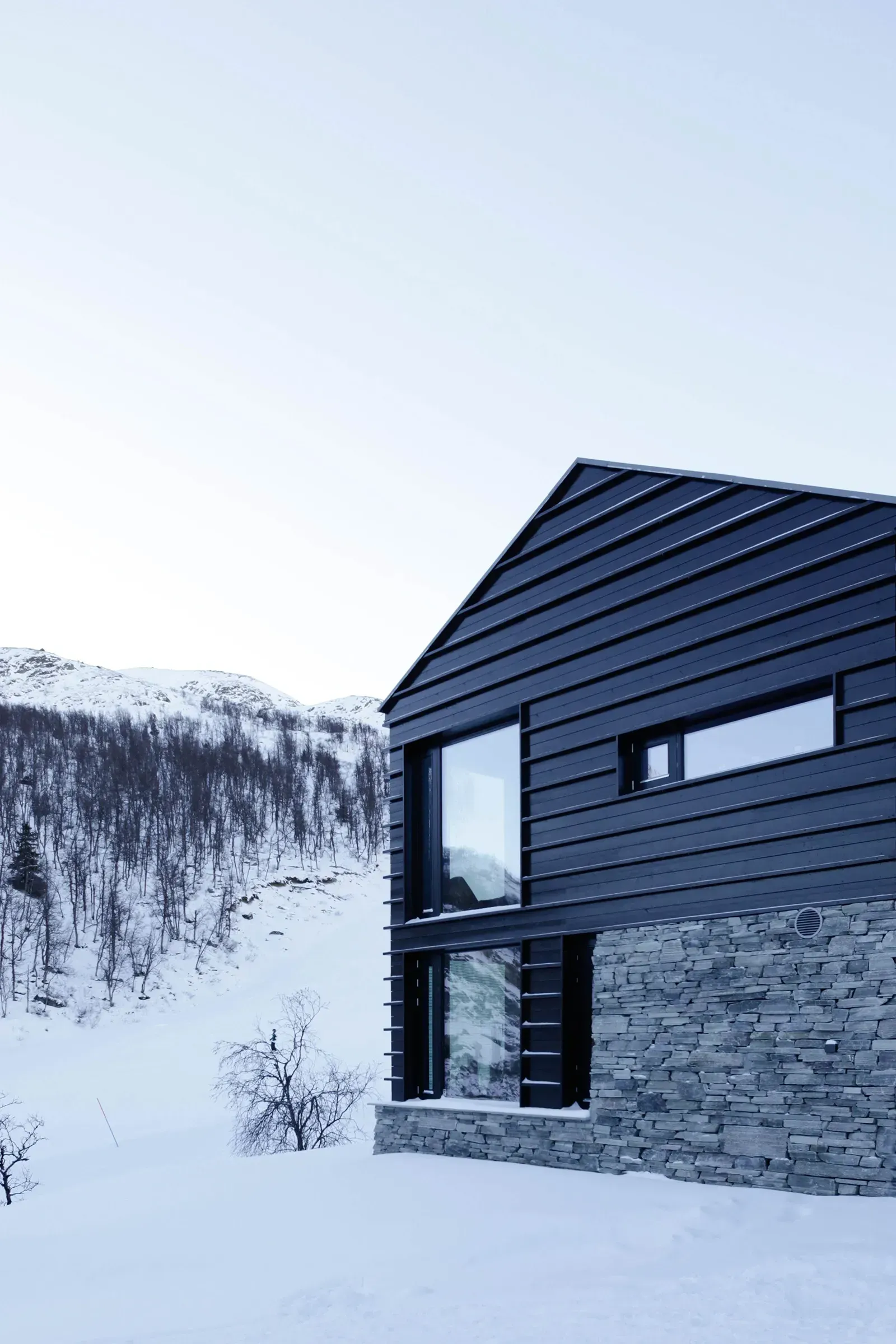Gavl med horisontale mørke trekledningsprofiler og store hjørnevinduer, fundamentert på naturstein.
