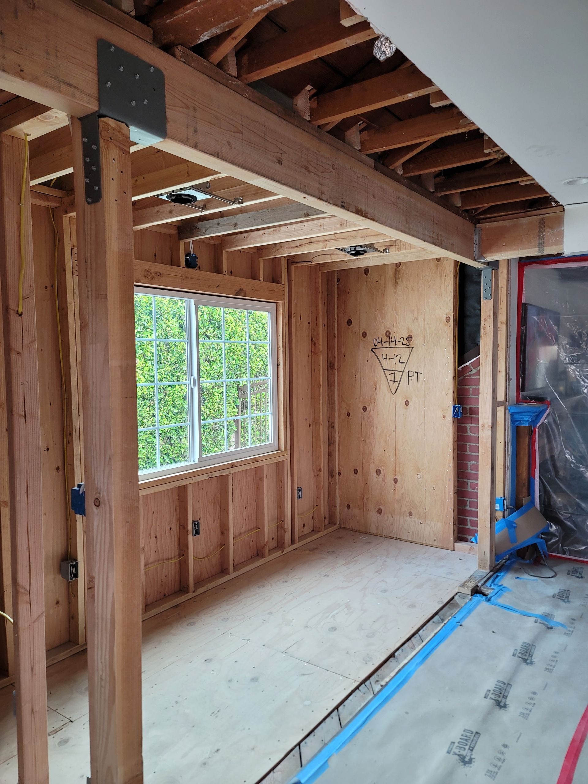 A room under renovation with exposed wooden wall studs, floor joists, a support beam, and a window.